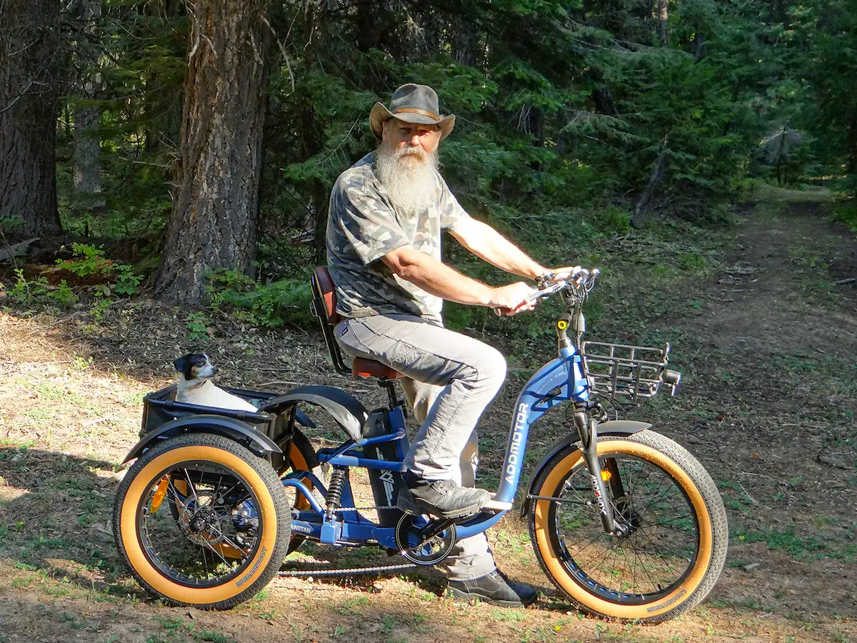 An old man takes his dog outdoors on an upright tricycle.